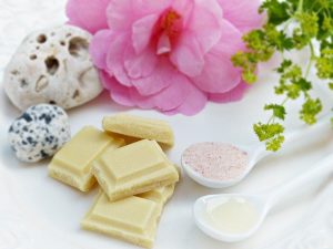 Pieces of white chocolate next to pink flower and spoons full of cocoa butter